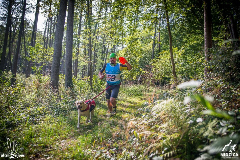 A man with a blue top and a green swim cap running through woods with his dog in a Swimrun race.