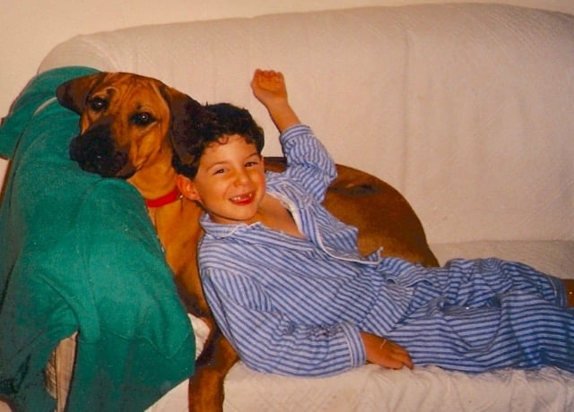 A young boy lying on a sofa wearing pyjamas, leaning against a Rhodesian Ridgeback